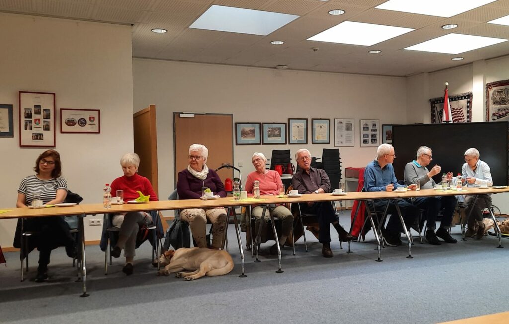 Am gedeckten Tisch sitzend Mitglieder der Gruppenversammlung, unter dem Tisch schläft Blindenführhund Ole
