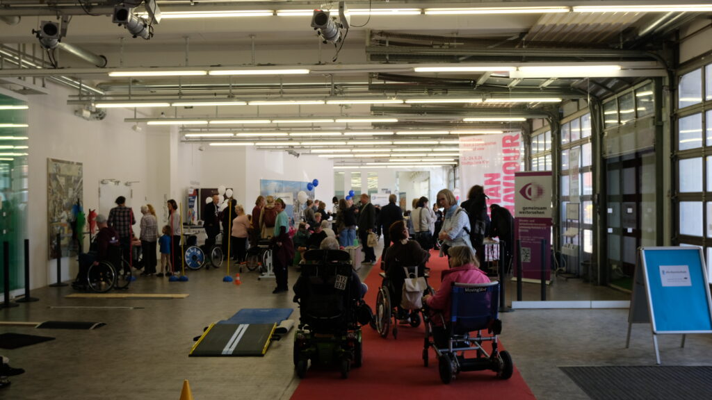 Viele Besucher sind im Foyer des Kulturforums Kiel zu sehen. Im Vordergrund mehrere Personen im Rollstuhl, die sich unterhalten und ein Rollstuhl-Parcours