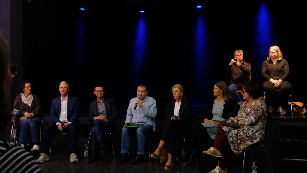 Sascha Lang moderiert die Gesprächsrunde und Live-Podcast Aufzeichnung zum Thema "Arbeit für alle?". Im Vordergrund sitzen Sascha Lang und seine Gesprächsgäste, im Hintergrund sind die Gebärdendolmetscherinnen zu sehen. 