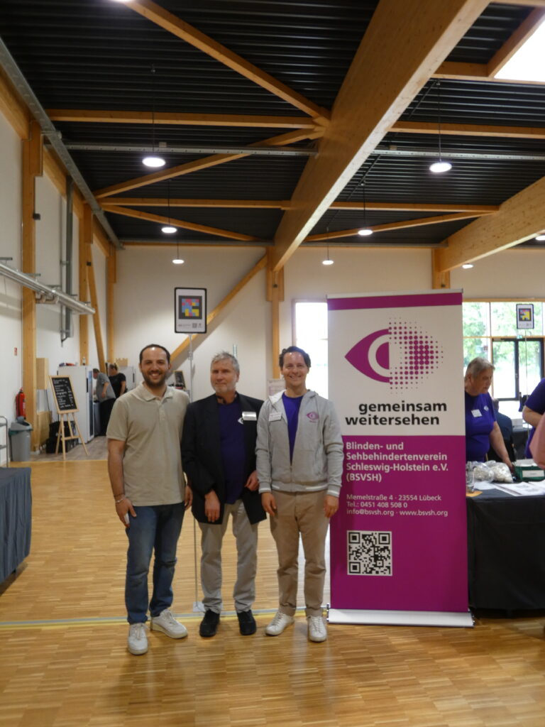 Nicola Cersullo (Vertreter von NaviLens in Deutschland), Vorstand Dr. Jürgen Trinkus und Geschäftsführer Martin Galeano stehen neben dem BSVSH Stand und Logo. Im Hintergrund ist ein NaviLens Code zu sehen.