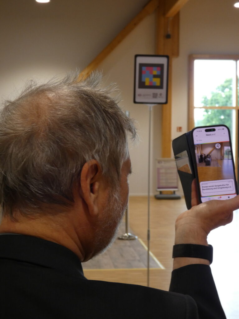 Dr. Jürgen Trinkus demonstriert die Nutzung der NaviLens App. Auf seinem Handybildschirm wird der Inhalt des vor ihm stehenden Codes angezeigt.