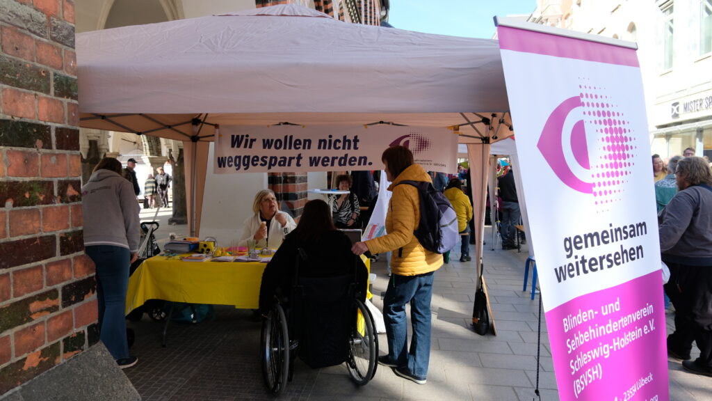 Karin Greiner berät Interessierte am BSVSH Infostand. Im Vordergrund steht ein Roll-Up mit dem BSVSH Logo und Slogan "gemeinsam weitersehen". Im Hintergrund hängt ein Banner mit der Aussage "Wir wollen nicht weggespart werden."