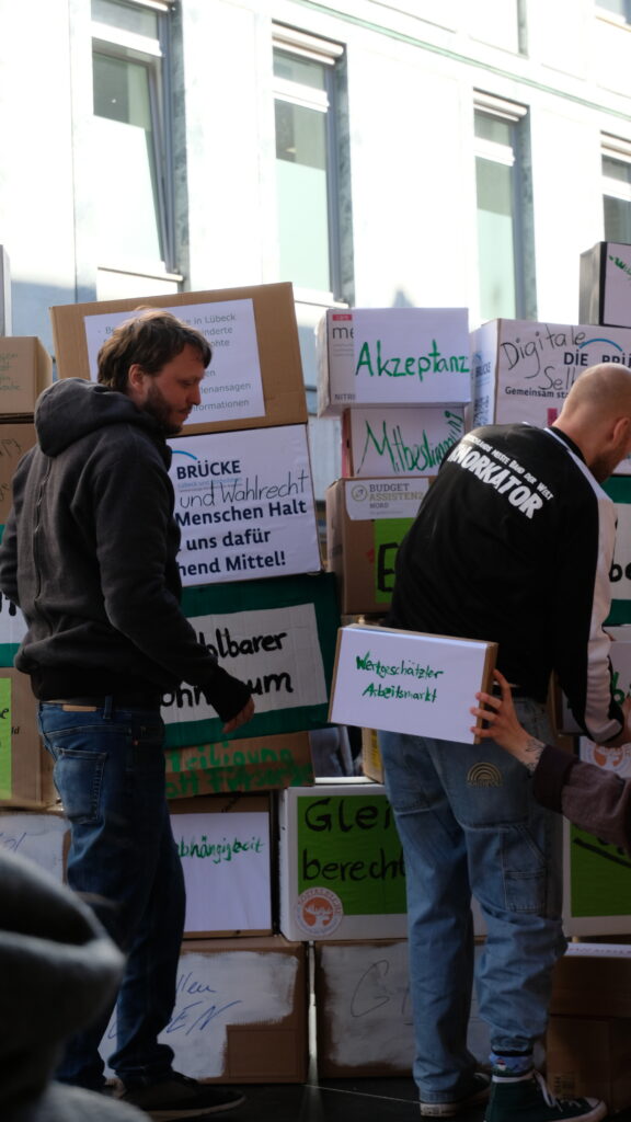 Freiwillige bauen eine Mauer aus Kartons auf auf denen Punkte notiert sind, die bei Inklusion aktuell gut und schlecht laufen. Von rechts wird ein Karton mit der Aufschrift "Wertschätzender Arbeitsmarkt" ins Bild gegeben. Im Hintergrund kann man z.B. lesen: Akzeptanz, Mitbestimmung und Digitale Selbstbestimmung