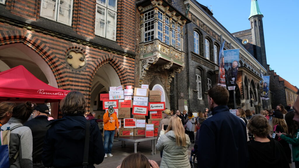 Ein Redner mit einem Megafon steht vor dem Lübecker Rathaus vor einer Mauer mit Kartons auf denen Beispiele für Barrieren und noch nicht funktionierende Inklusion vermerkt sind. Man kann z.B. lesen: Fremdbestimmung, teurer Wohnraum, Diskriminierung und Intoleranz
