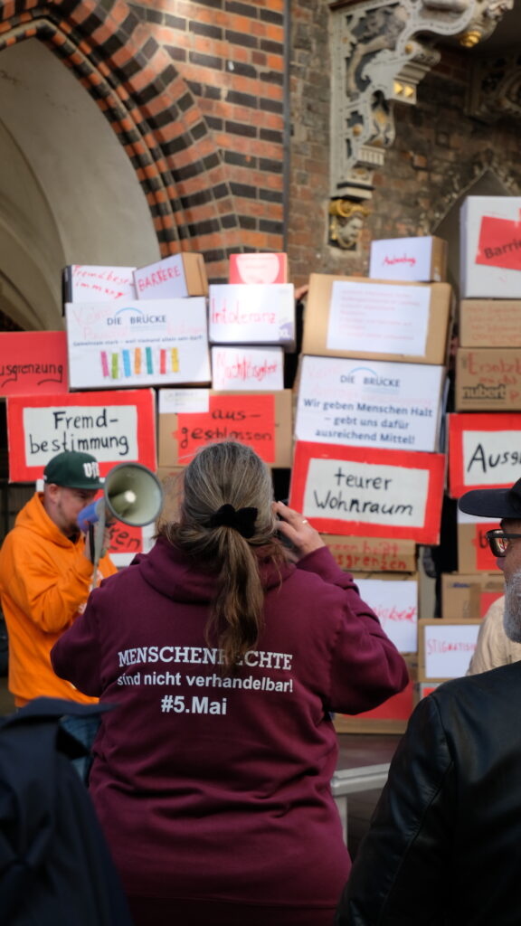 Vor einer Mauer mit Beispielen für Barrieren und noch nicht funktionierender Inklusion steht eine Person mit einem Pullover mit der Aufschrift "MENSCHENRECHTE sind nicht verhandelbar! #5. Mai"