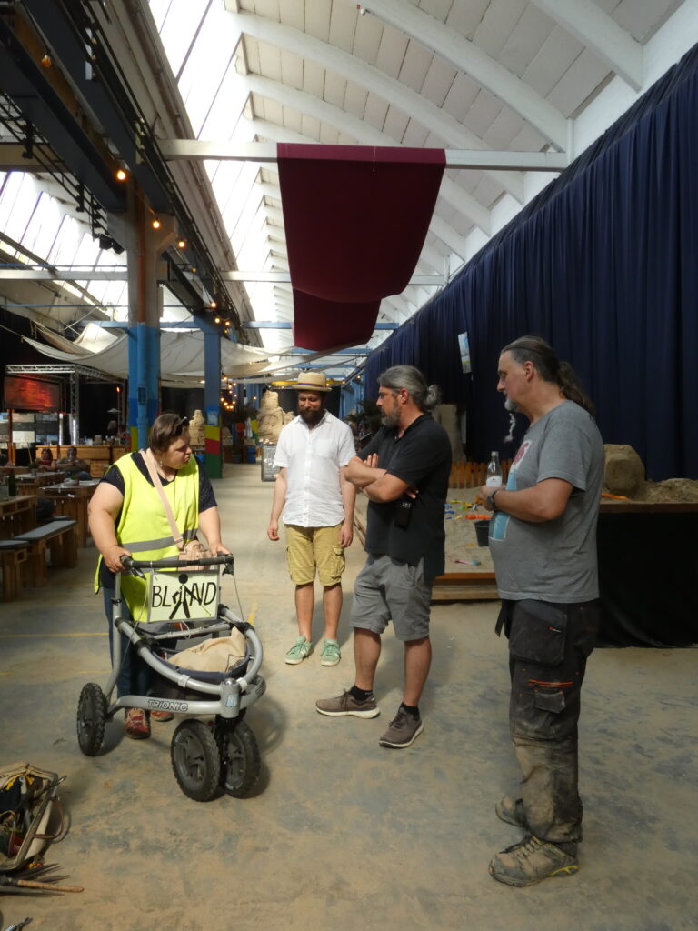 Teilnehmer, Künstler und Ausstellungsleiter im Gespräch während der Pause. Vier Personen in einer Ausstellungshalle im Gespräch. Eine Frau mit gelber Warnweste stützt sich auf einen Rollator mit der Aufschrift „BLIND".
