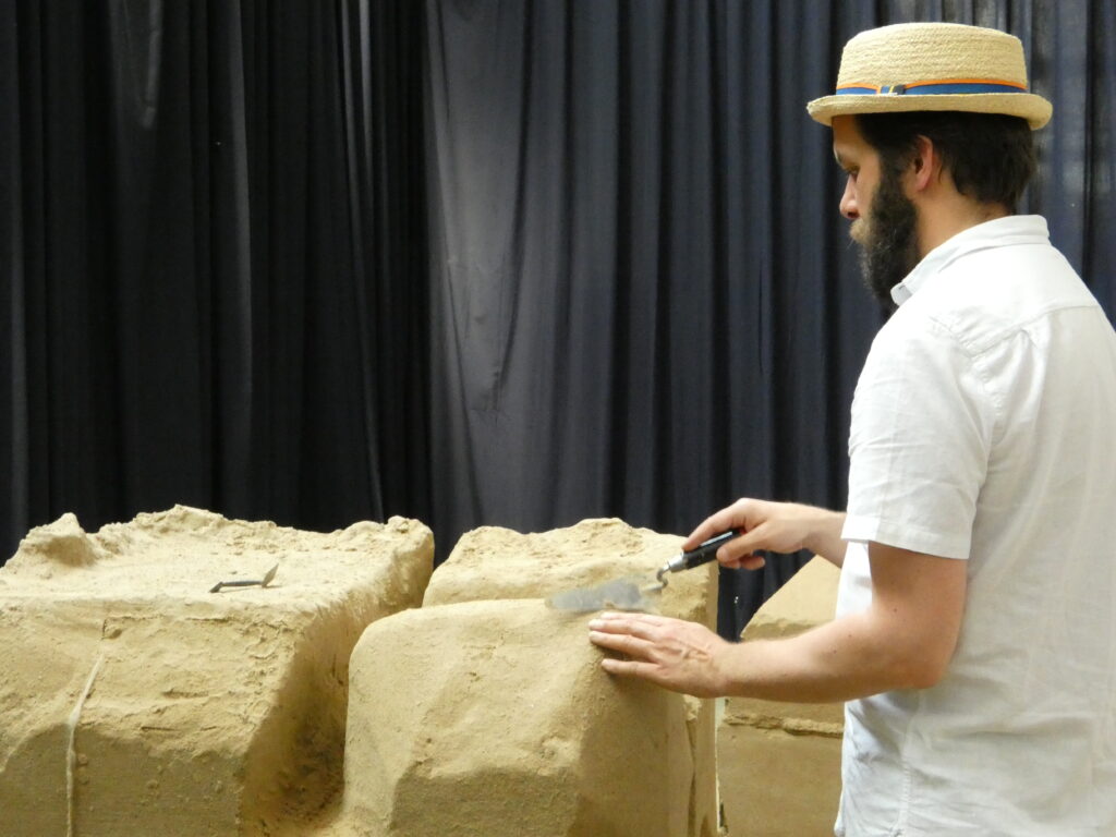 Jordan Smith ertastet die Form seiner Skulptur und bearbeitet mit einem Werkzeug die Kante des großen Sandblocks.