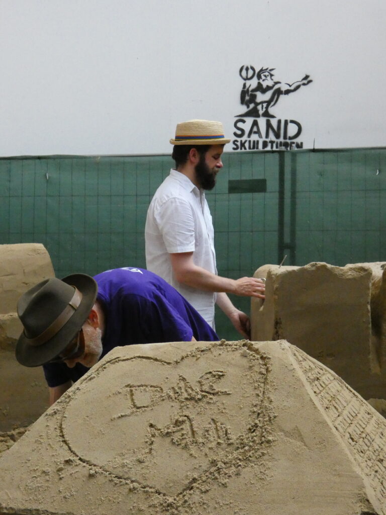 Zwei Teilnehmer bearbeiten gleichzeitig Sandskulpturen in einer Halle; im Hintergrund das aufgesprühte Logo „Sand Skulpturen".