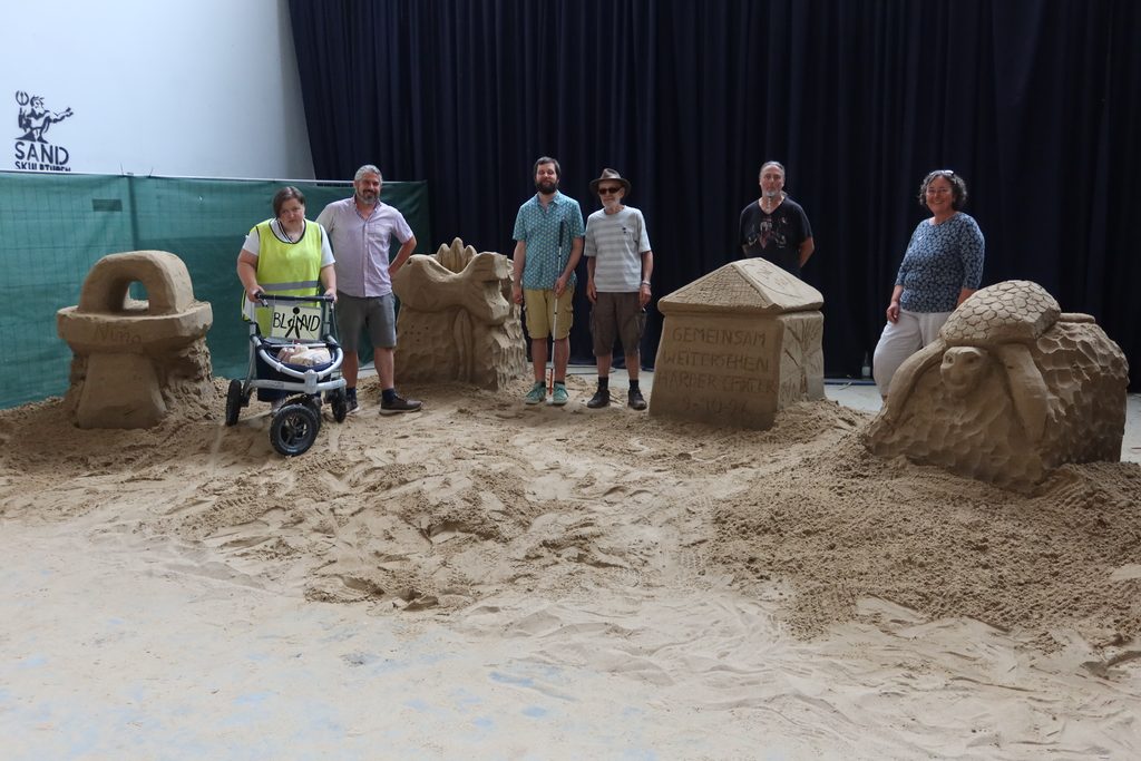 Die vier Teilnehmenden stehen neben ihren Kunstwerken und lachen in die Kamera. Ausstellungsleiter Oliver Hartmann und Sandkünstler Martin De Zoete sind mit auf dem Gruppenbild