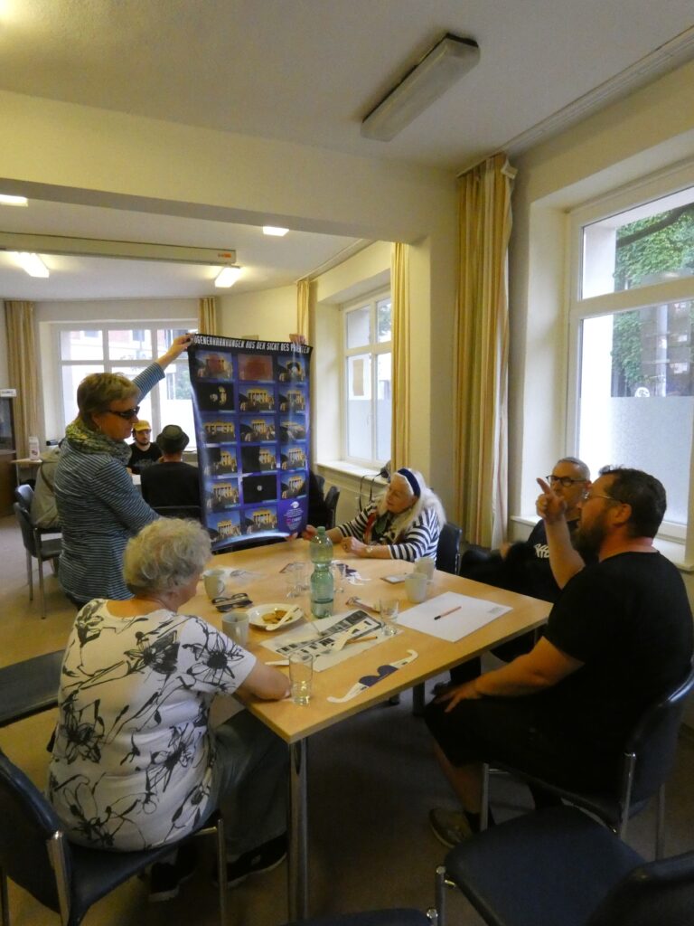 Eine Person präsentiert einer Gruppe ein bebildertes Plakat mit dem Titel "Augenerkrankungen aus Sicht des Patienten". Die Teilnehmenden sitzen an Tischen und diskutieren.