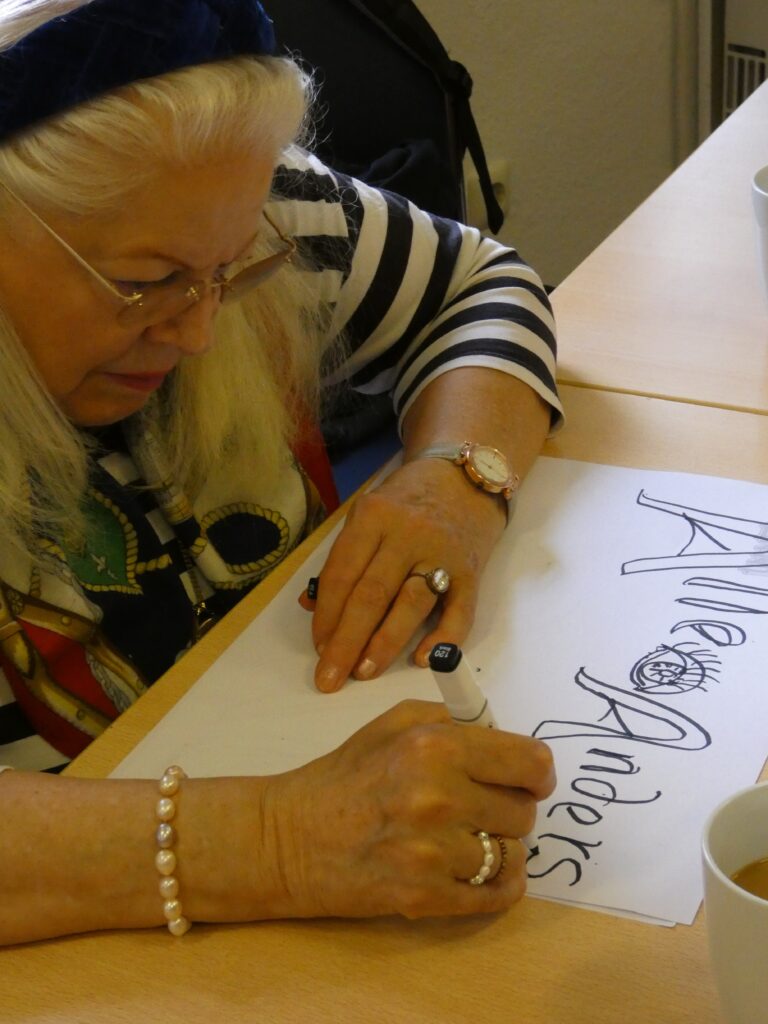 Eine Frau zeichnet mit einem Stift in geschwungenen Buchstaben "Alle sehen anders" auf ein großes Blatt Papier.