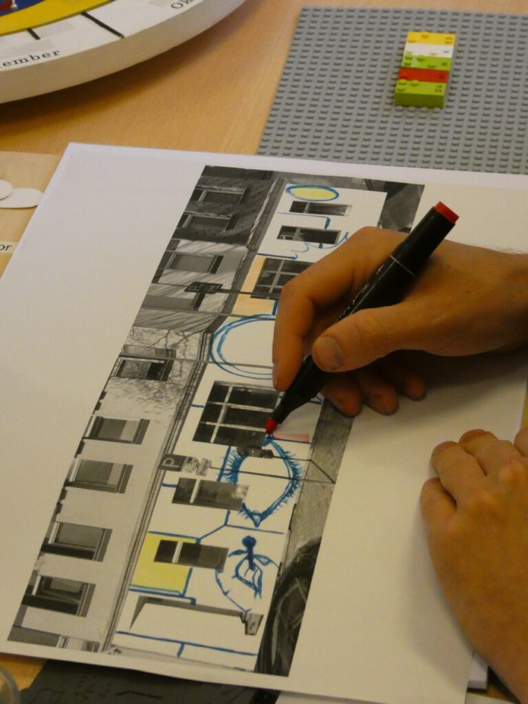 Eine Hand gestaltet mit einem schwarzen und roten Marker ein Schwarz-Weiß-Foto der Fassade mit blauen Zeichnungen.