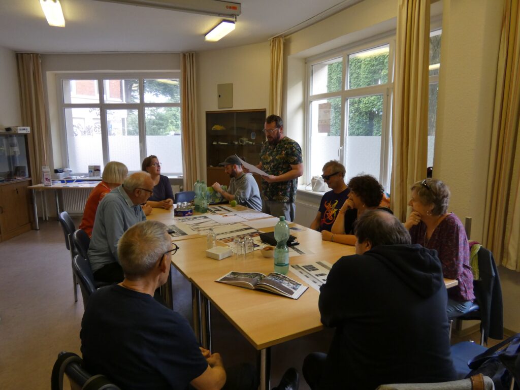 Eine Gruppe sitzt um einen Tisch, während ein Mann im Stehen Ergebnisse des Workshops vorstellt.
