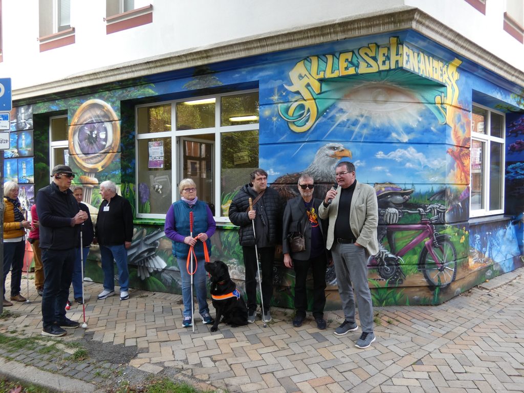Bürgermeister Gerwin Stöcken hält ein Grußwort zur Einweihung der Fassade und steht mit der BSVSH Regionalgruppe Kiel unter dem Schriftzug "Alle Sehen Anders" auf der bunten Fassade der Mela31